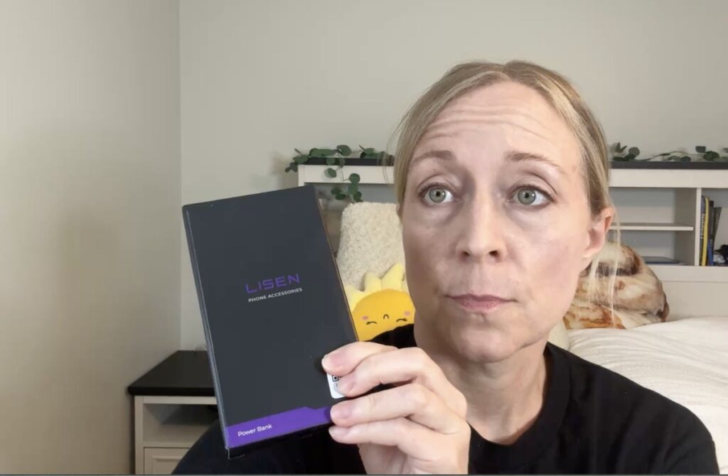 Jen holds up a black Lisen Phone Accessories power bank box with a purple accent stripe in a bedroom.