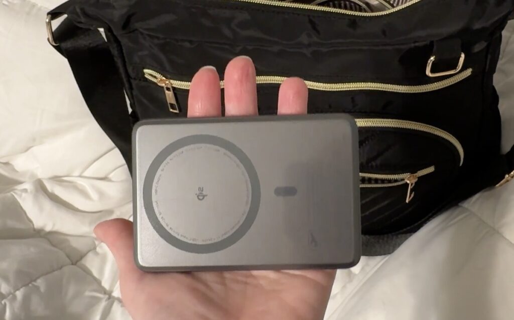 Hand holding a slim gray Qi wireless power bank in front of a black nylon bag with gold zippers.