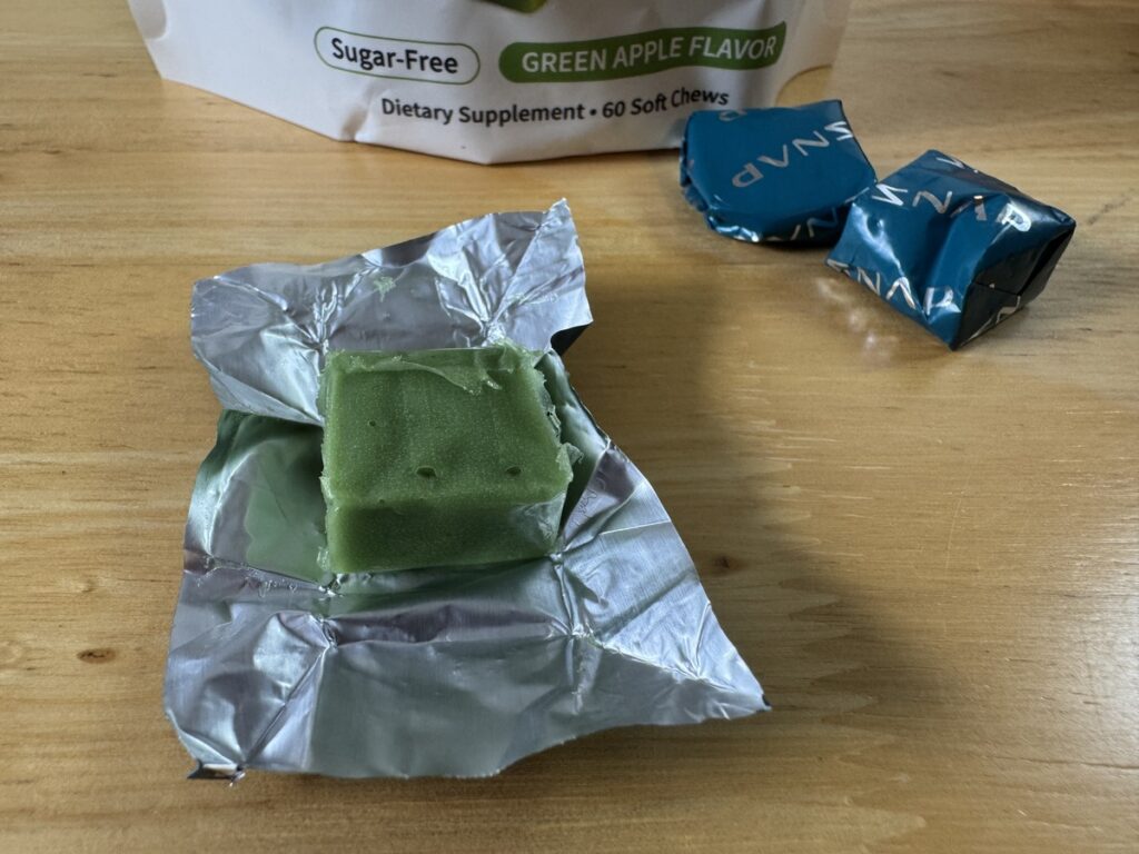 Unwrapped green apple SNAP soft chew on foil next to two blue-wrapped chews and the sugar-free supplement pouch.