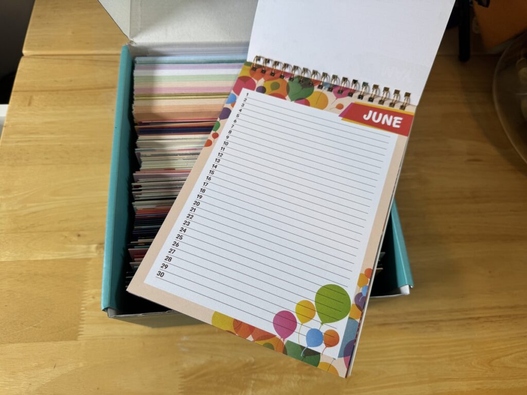 Spiral-bound monthly planner open to the June page with colorful dot decoration, sitting on a greeting card box.