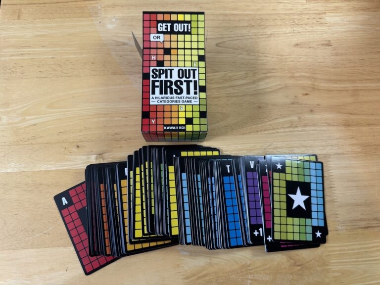 Spit Out First card game by Kawaii Koi with pixel-grid box and a fanned pile of colorful category cards on a wood table.