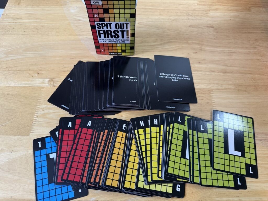 Spit Out First card game spread on wood floor with black cards and colorful grid-letter cards in green, yellow, red, blue.