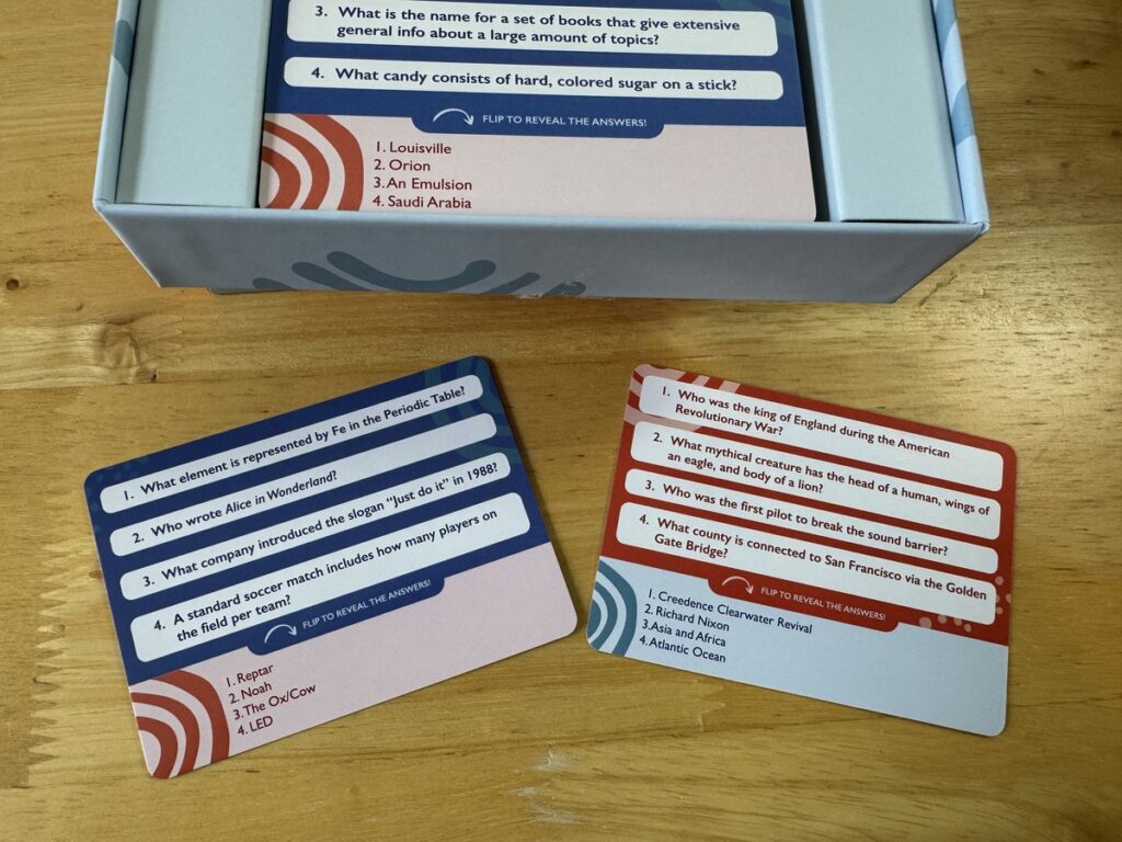 Trivia card game box with red and blue question cards fanned out on a light wood tabletop.