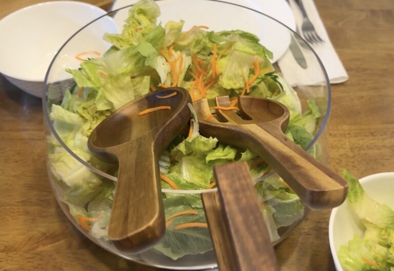 Two wooden salad server hands resting on iceberg lettuce and shredded carrot in a clear glass bowl on a wood table.