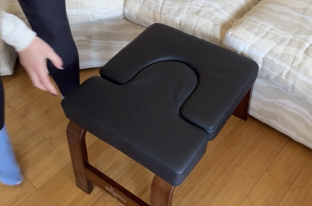 Black padded yoga headstand inversion bench with U-shaped cutout and wooden legs on a hardwood floor beside a beige sofa.
