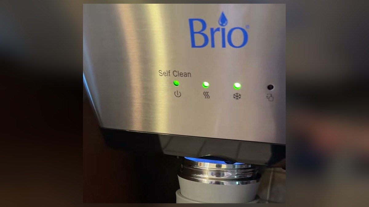 Close-up of the Brio water dispenser front panel with the Self Clean, power, hot, and cold indicator lights and a tall tumbler being placed under the spout
