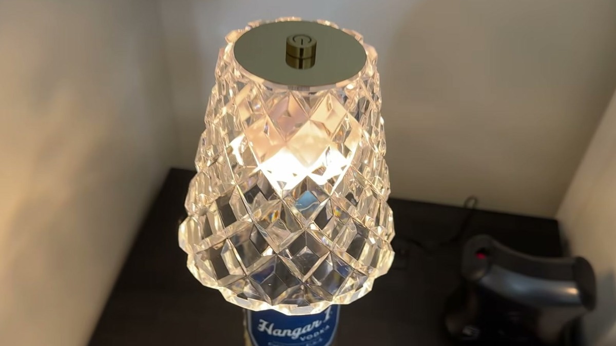 Jiofaviu wine bottle lamp lit in warm white on a Hangar 1 vodka bottle, its faceted clear acrylic shade and round gold touch plate glowing on a dark wood nightstand
