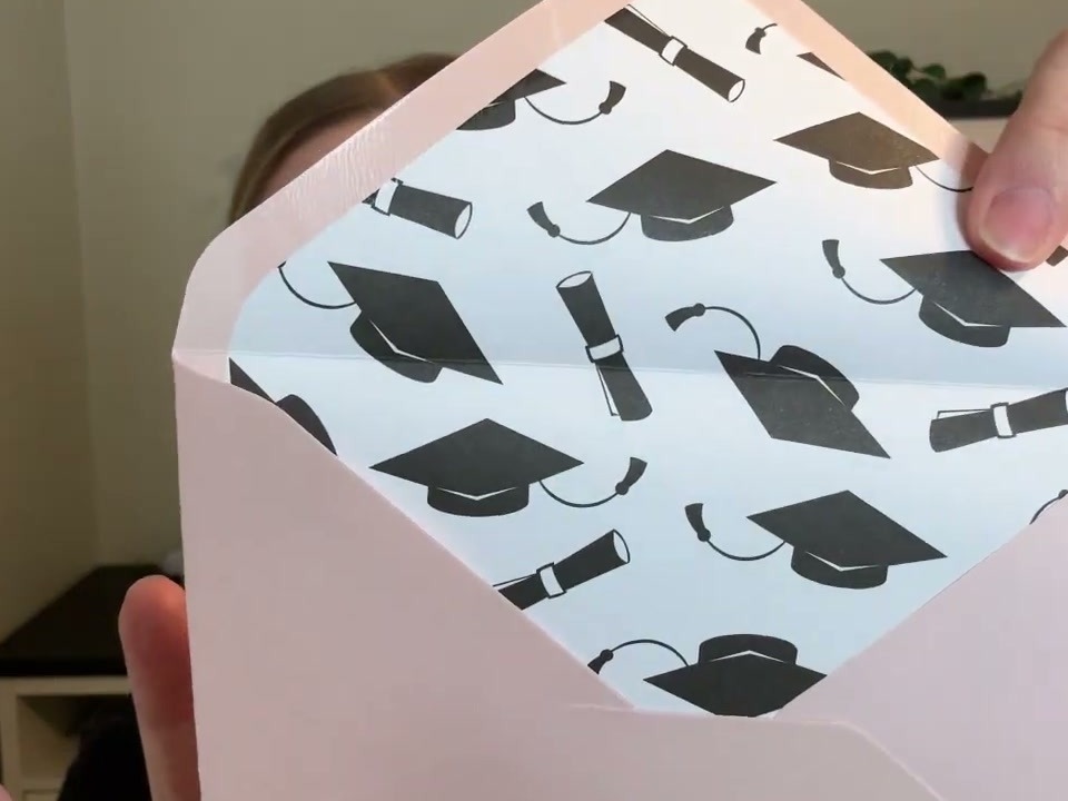 A blush pink Decorably graduation card envelope opened to show the printed graduation cap lining inside