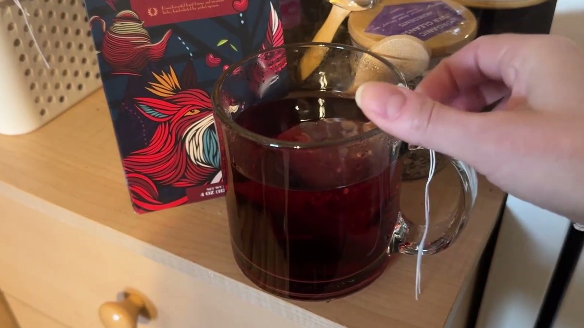Glass mug of deep red brewed ENZO Crimson Harvest tea on a dresser with the box behind it and a hand lifting the tea bag string