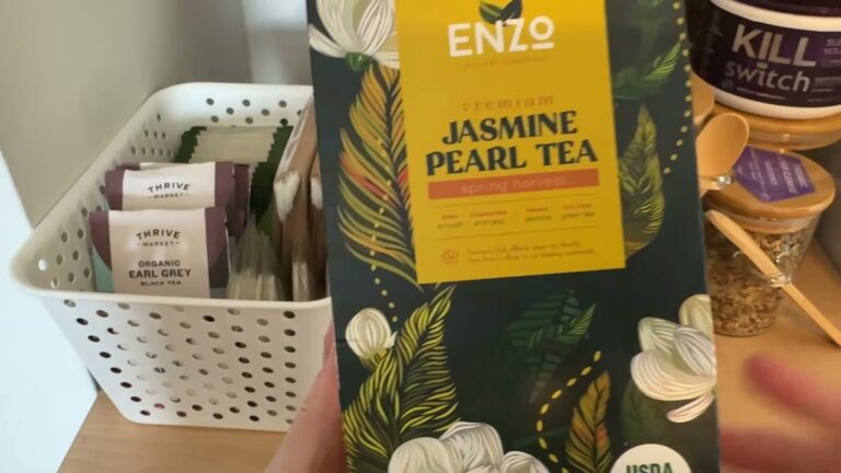 Enzo Dragon Pearl jasmine tea box, dark green with yellow label and white jasmine flowers, standing on a counter next to a glass mug and a basket of tea bags