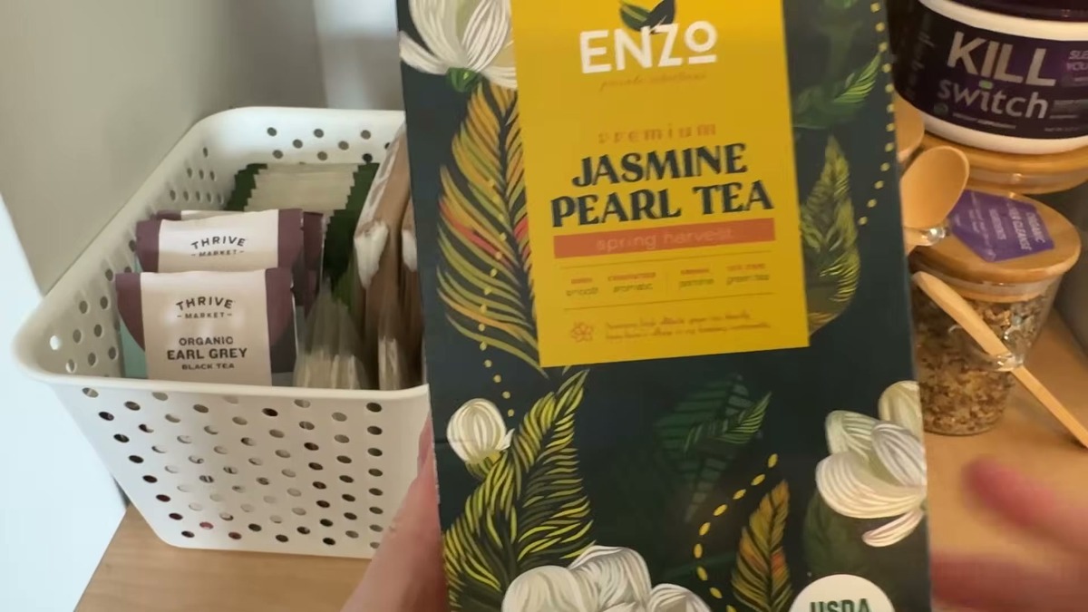 Enzo Dragon Pearl jasmine tea box, dark green with yellow label and white jasmine flowers, standing on a counter next to a glass mug and a basket of tea bags