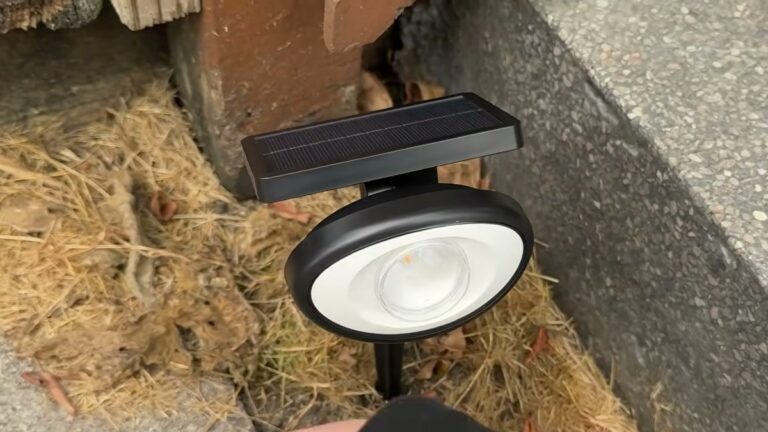 Black Fuligent solar landscape spotlight staked in dry mulch beside a concrete deck step, with its solar panel angled on top of the round light head