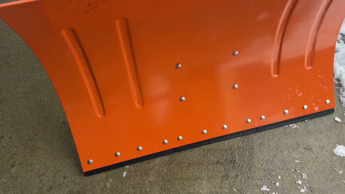Front of the Garvee wheeled snow shovel's orange 30 inch carbon steel blade showing the rivets and the black protective strip along the bottom edge