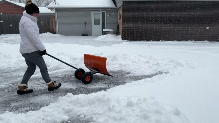 Garvee 30 inch wheeled snow shovel with an orange carbon steel blade and 10 inch rubber wheels being pushed across a snowy paved driveway