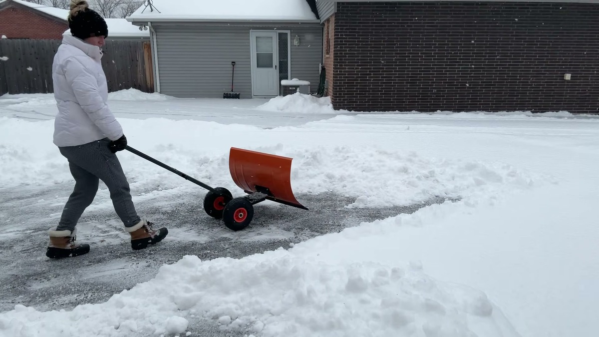 Garvee 30 inch wheeled snow shovel with an orange carbon steel blade and 10 inch rubber wheels being pushed across a snowy paved driveway