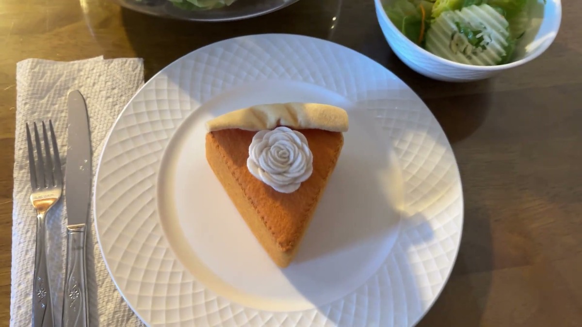 A single Hahafelt felt pumpkin pie slice with a cream felt crust and white whipped cream rosette sitting on a white dinner plate next to a fork and knife
