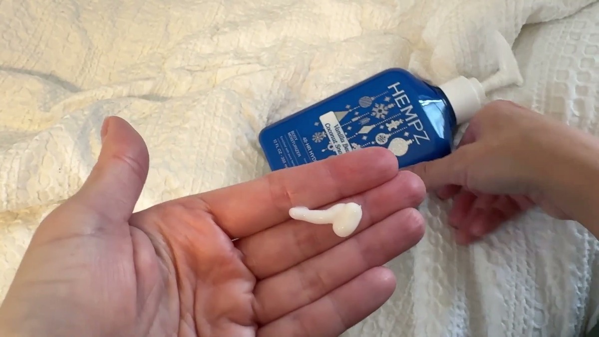 A dollop of white Hempz Vanilla Bean and Coconut Snowball lotion on an open palm, with the blue bottle lying on a white blanket behind it