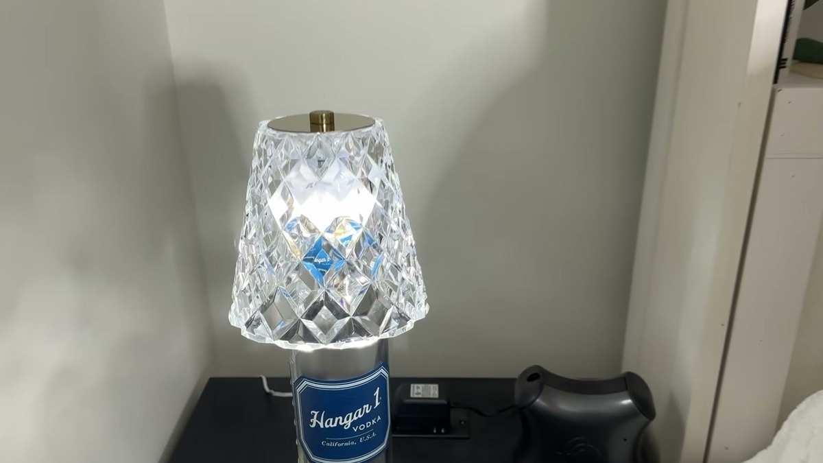The Jiofaviu wine bottle lamp switched to its cool white setting, the clear faceted acrylic shade seated on a tall Hangar 1 vodka bottle on a bedroom nightstand