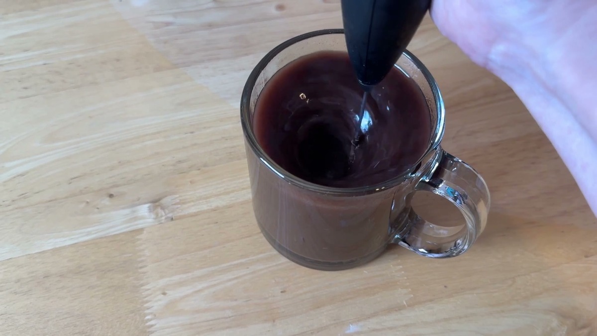 A handheld frother mixing Killswitch hot cocoa in a clear glass mug on a wooden counter