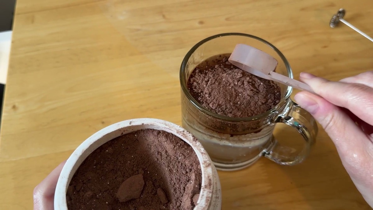 A scoop of Killswitch cocoa powder going into a clear glass mug next to the open canister