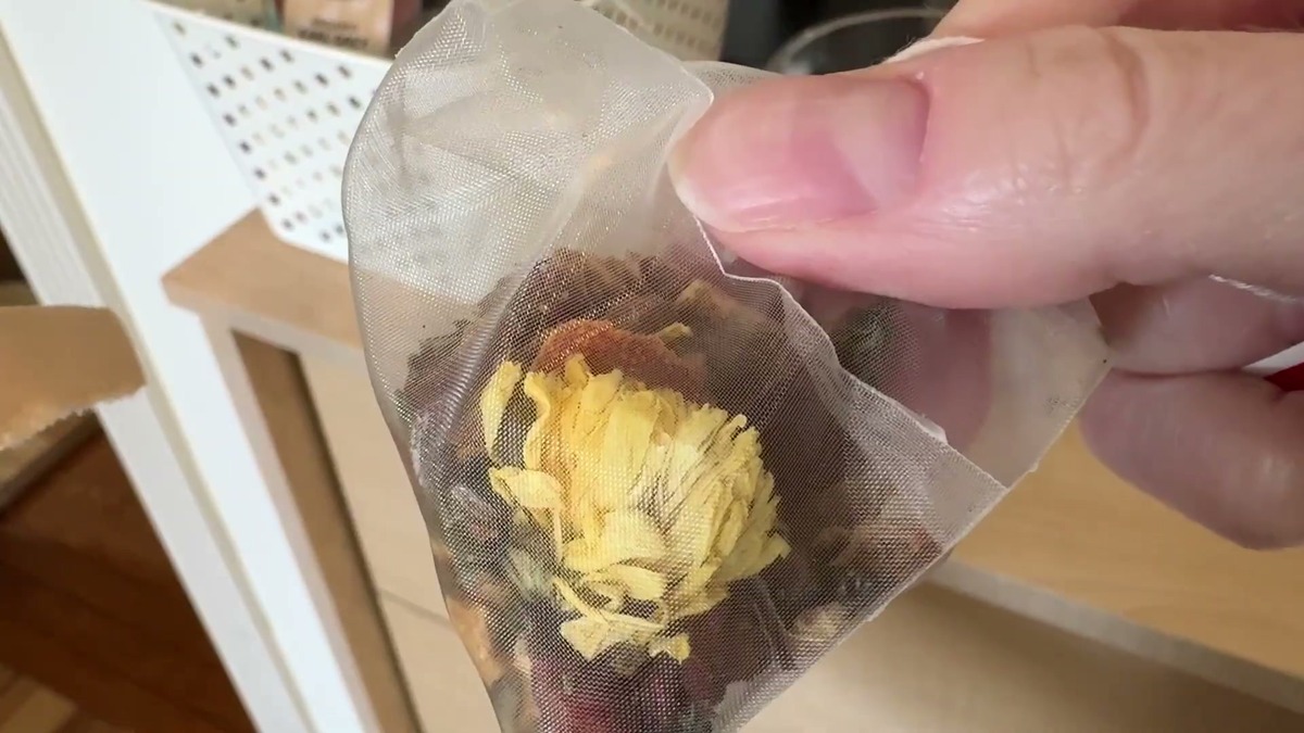 Pyramid tea bag of liver and kidney detox tea held up to show a whole dried chrysanthemum flower and loose herbs inside