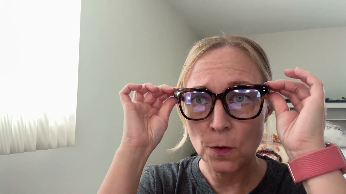 Jen wearing the miyya oversized tortoiseshell blue light glasses at her desk, adjusting them with both hands, faint blue reflection visible on the lenses