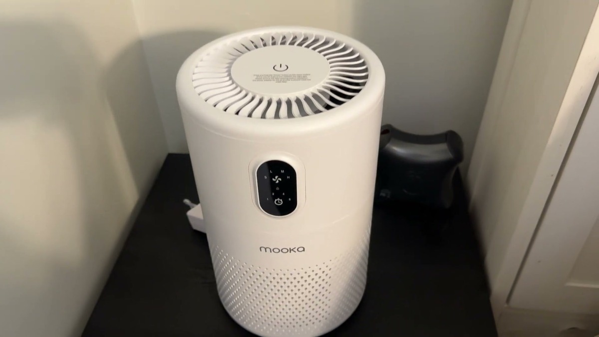 White MOOKA B-D02L air purifier sitting on a small black end table, with its round top fan grille and front touch control panel in view