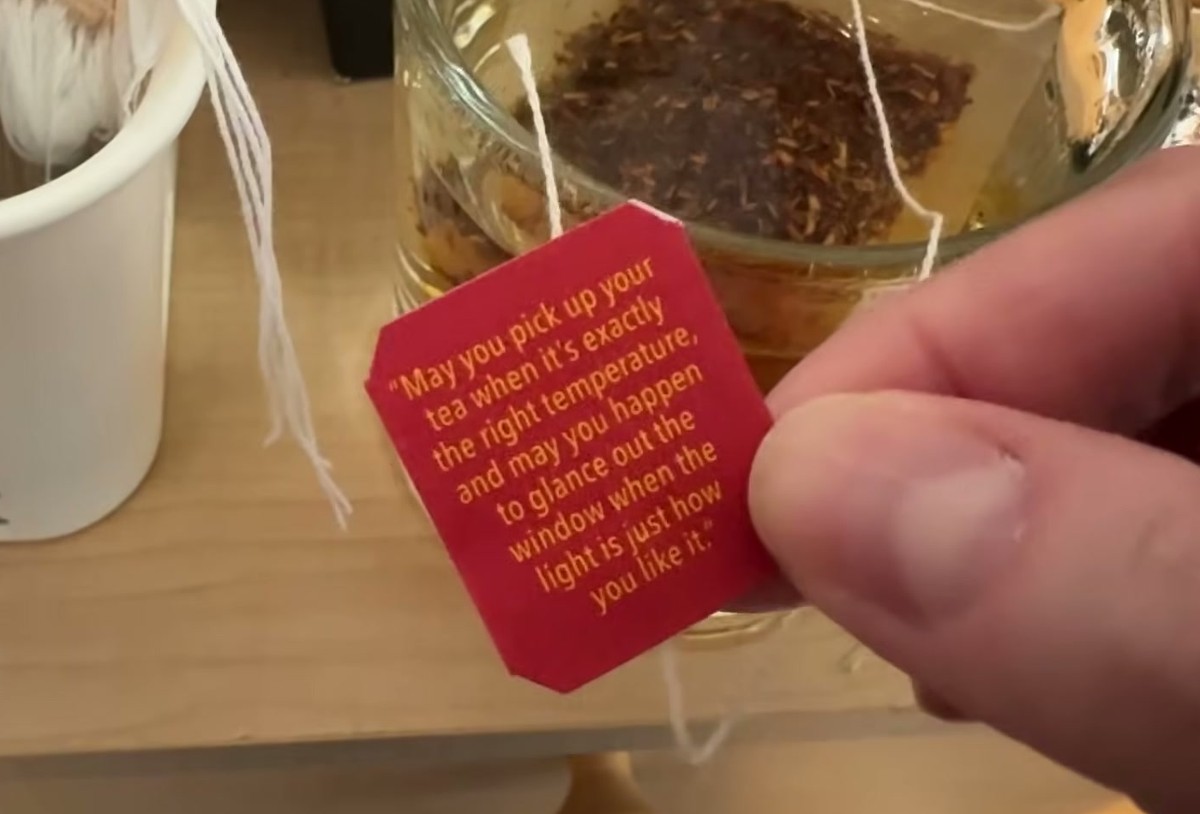 Red My Red Tea bag tag held over a steeping mug, printed with a quote about picking up your tea at exactly the right temperature and glancing out the window when the light is just how you like it