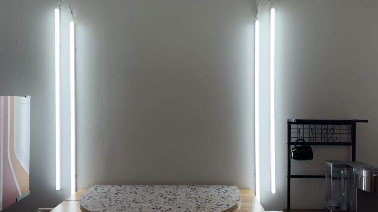 Four Pak 4FT LED shop lights mounted vertically in two linked pairs on a white office wall, switched on and lighting a terrazzo photo table.