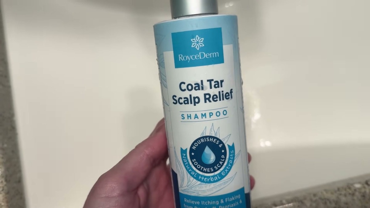 RoyceDerm Coal Tar Scalp Relief Shampoo bottle held head-on over a bathroom sink, white and blue label with the 2% coal tar extract line