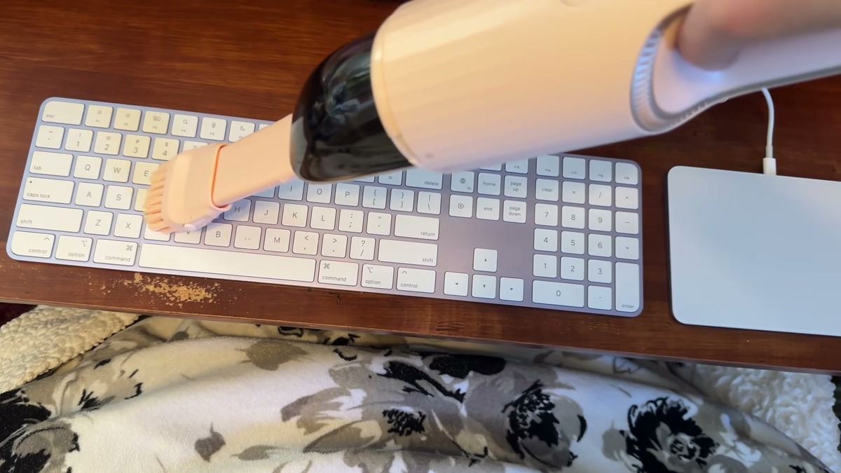 The pink STARUMENT hand vacuum with its brush attachment sweeping across a white keyboard, with crumbs scattered on the wooden desk beside it.