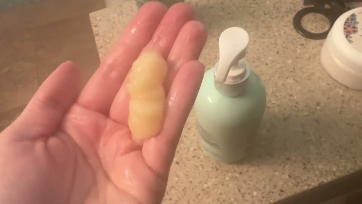 A dollop of pale yellow Svvimer rosemary conditioner in an open palm, with the mint green pump bottle on the counter behind it