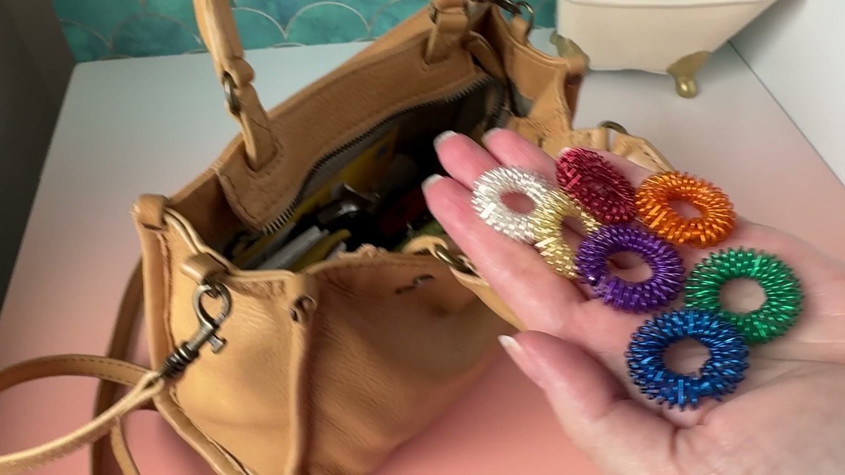 A hand holding seven colorful Tondiamo spiky sensory rings next to an open tan leather purse