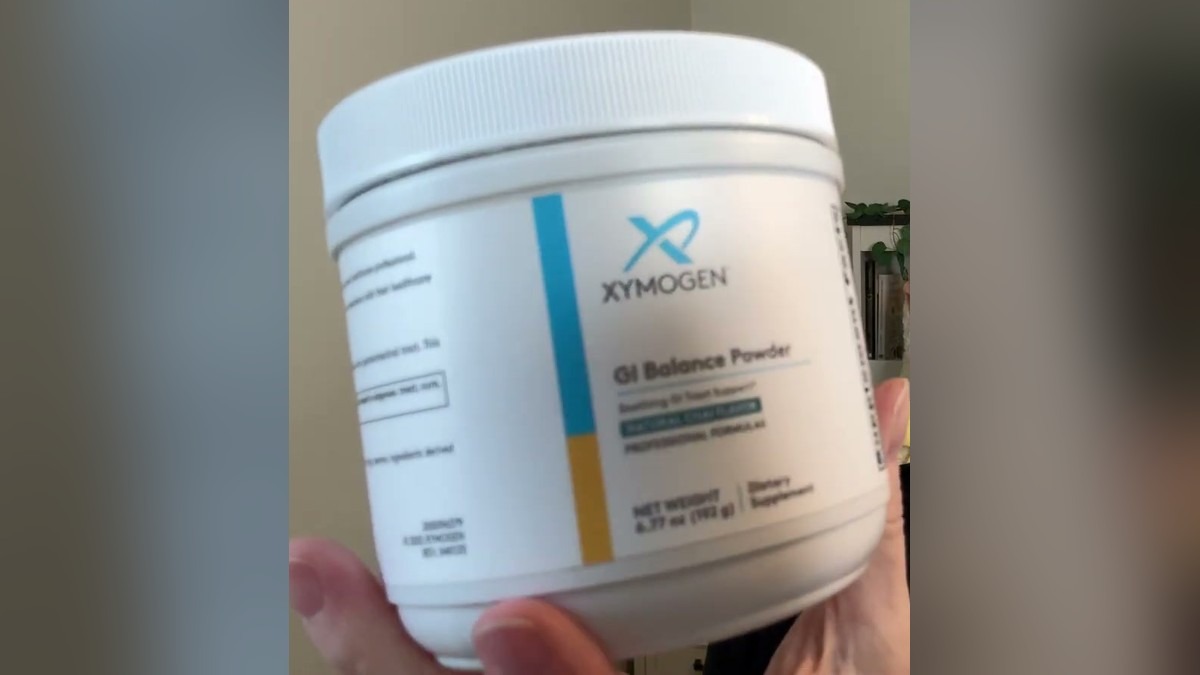XYMOGEN GI Balance Powder in natural chai flavor, a white 6.77 oz tub with a blue and gold label, held up in one hand