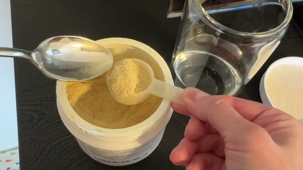 A scoop of tan XYMOGEN GI Balance chai powder being leveled with a spoon over the open tub, next to a glass of cold water