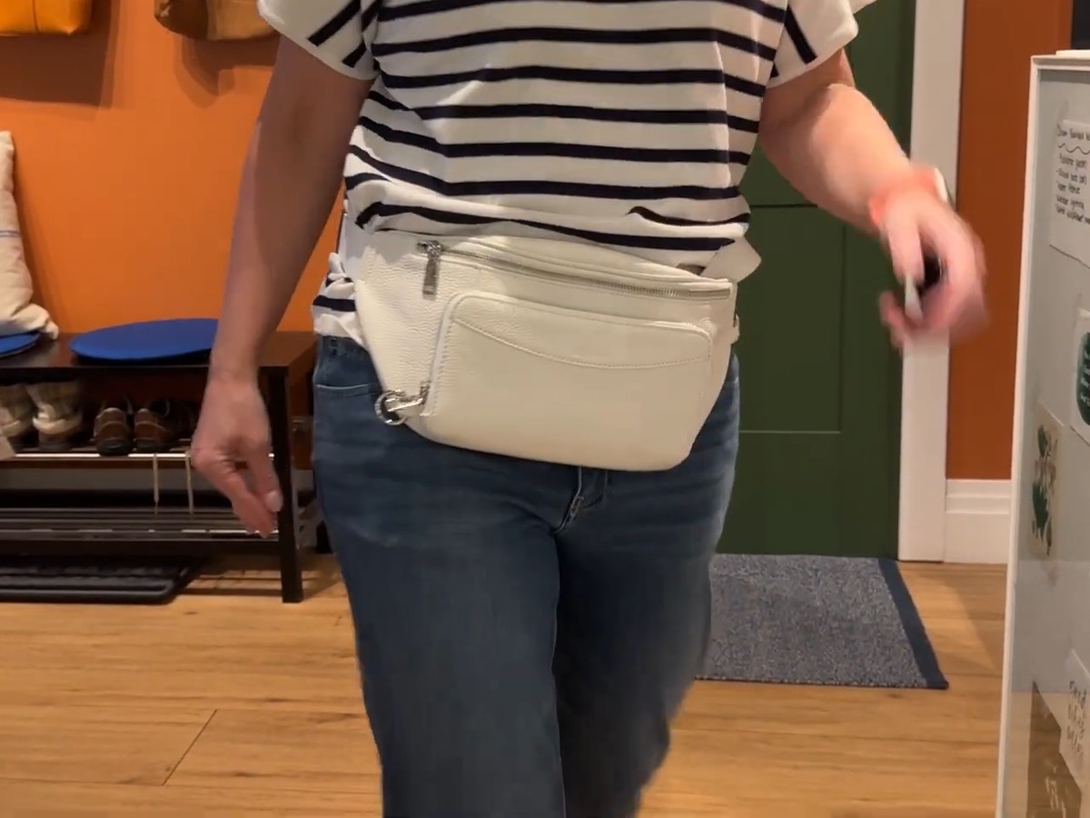 White befen leather fanny pack worn at the waist over jeans and a striped tee while walking through a furniture store.