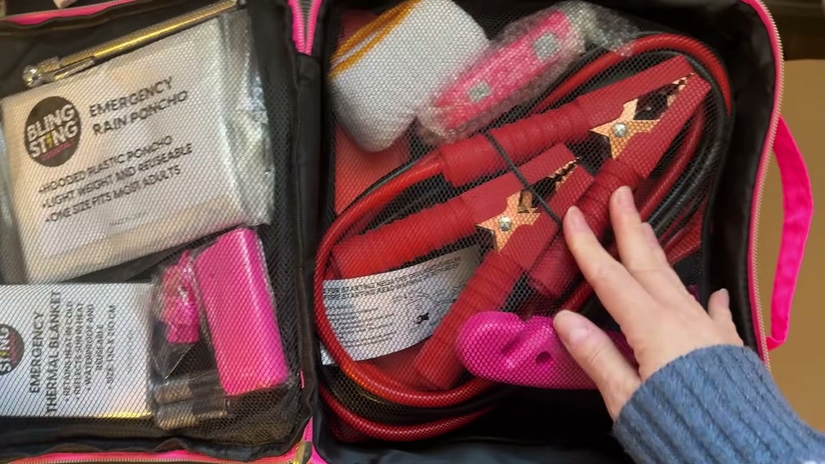 Open BLINGSTING car emergency kit showing red jumper cables behind a mesh net, a pink escape tool, a rain poncho, and an emergency thermal blanket