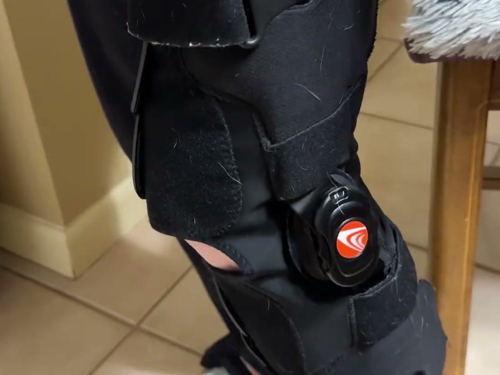 Close-up of the red Breg thumbwheel dial on the side hinge of the Freestyle OA knee brace, with the hook-and-loop straps wrapped around the leg