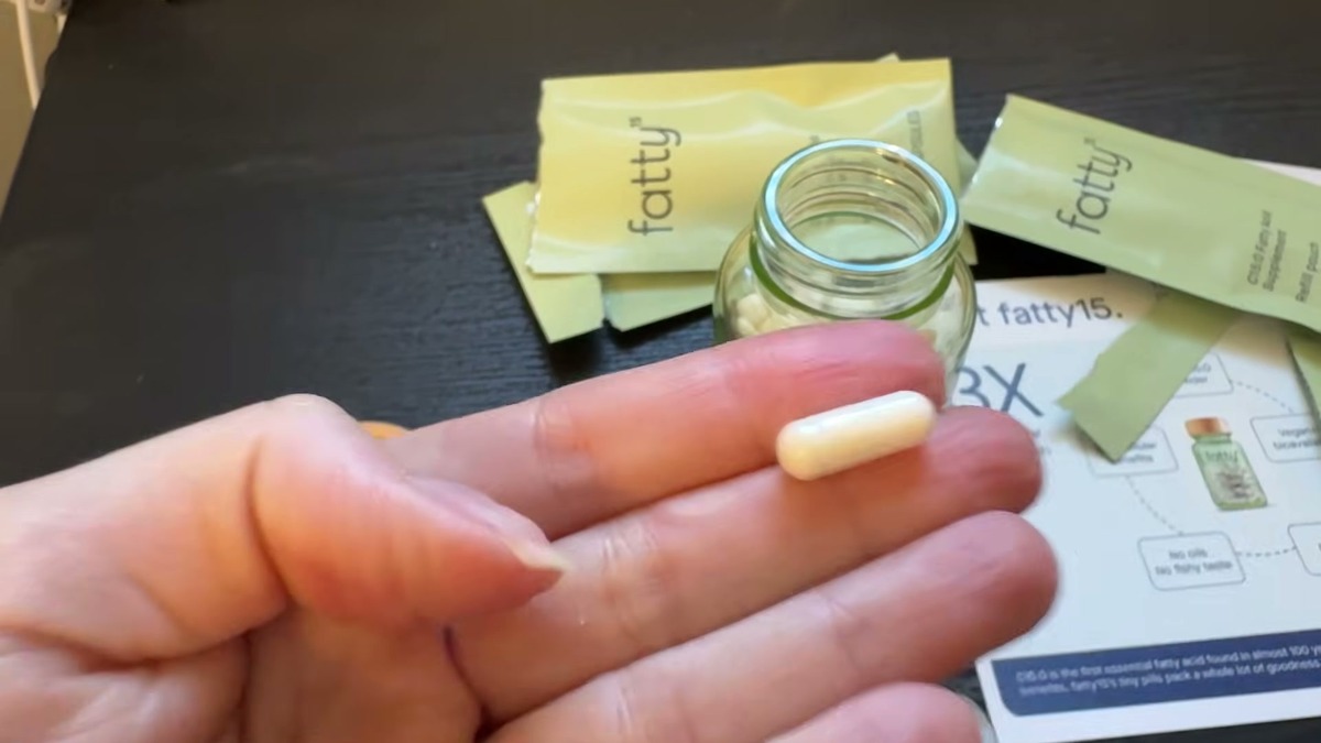 One small Fatty15 capsule resting on two fingers, with the open glass jar and refill pouches behind it