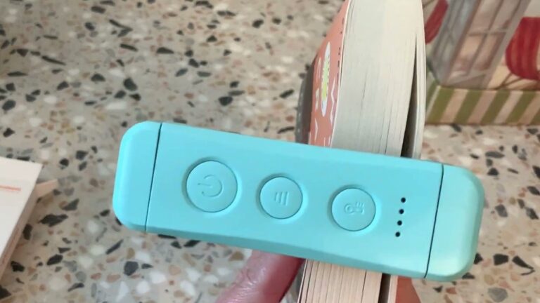 Teal Glocusent bookmark-style book light clipped to the back of a paperback, showing its three buttons and four battery indicator dots