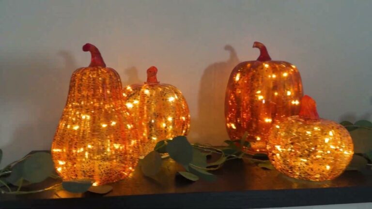 Four Glooglitter orange-gold glass pumpkins in different sizes glowing with fairy lights on a dark fireplace mantel with eucalyptus greenery