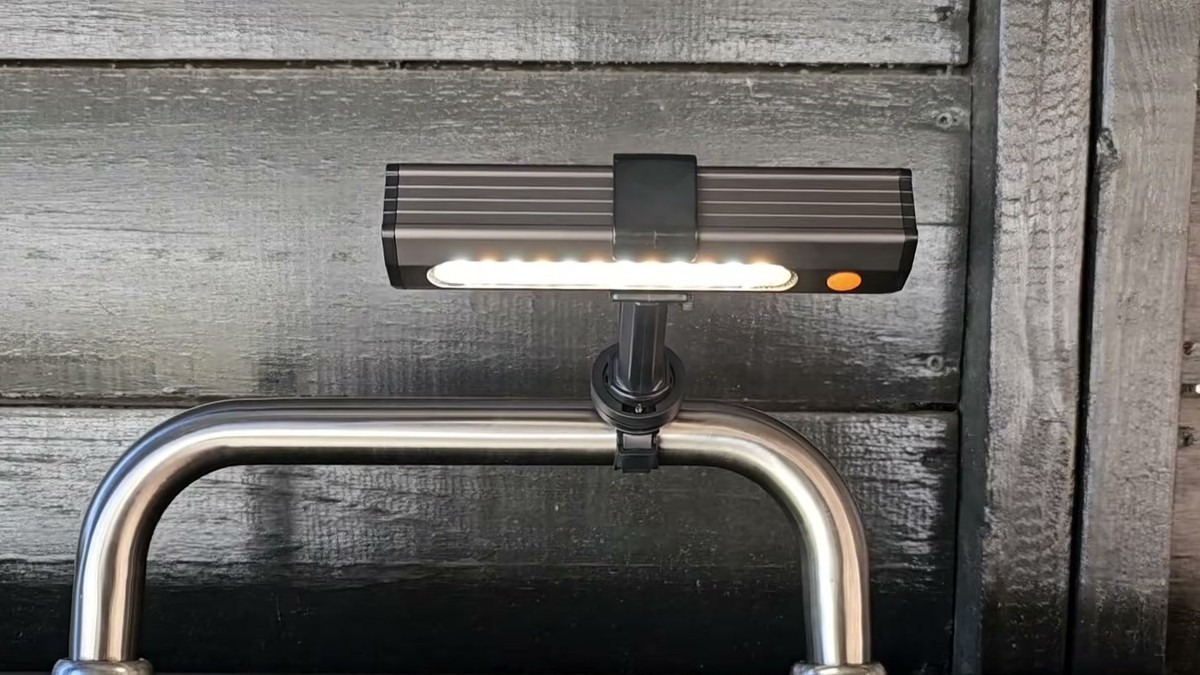 Coinicxcel LED grill light clamped to a stainless Blackstone griddle lid handle, lit up against a dark wood fence