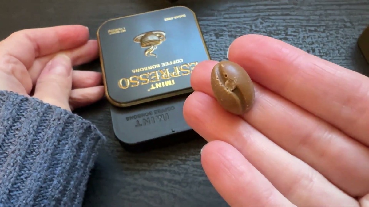 A single bean-shaped I'MINT coffee hard candy held in fingertips above the black Espresso tin