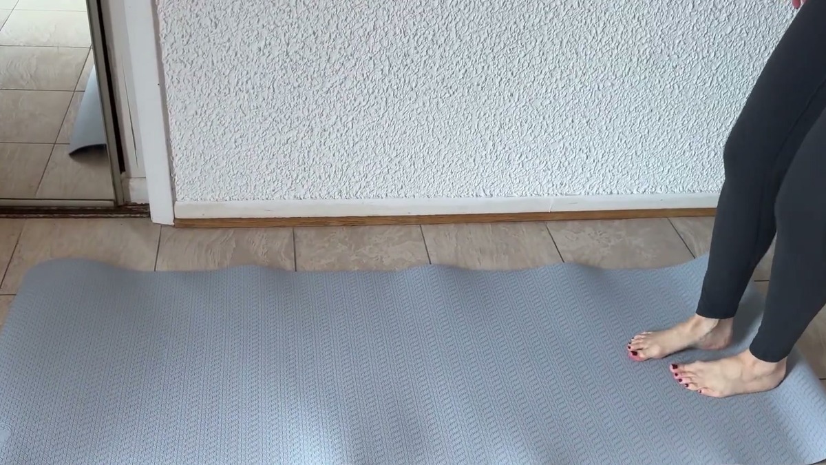 Light gray IUGA 6mm yoga mat unrolled on a tile floor, with bare feet standing at one end