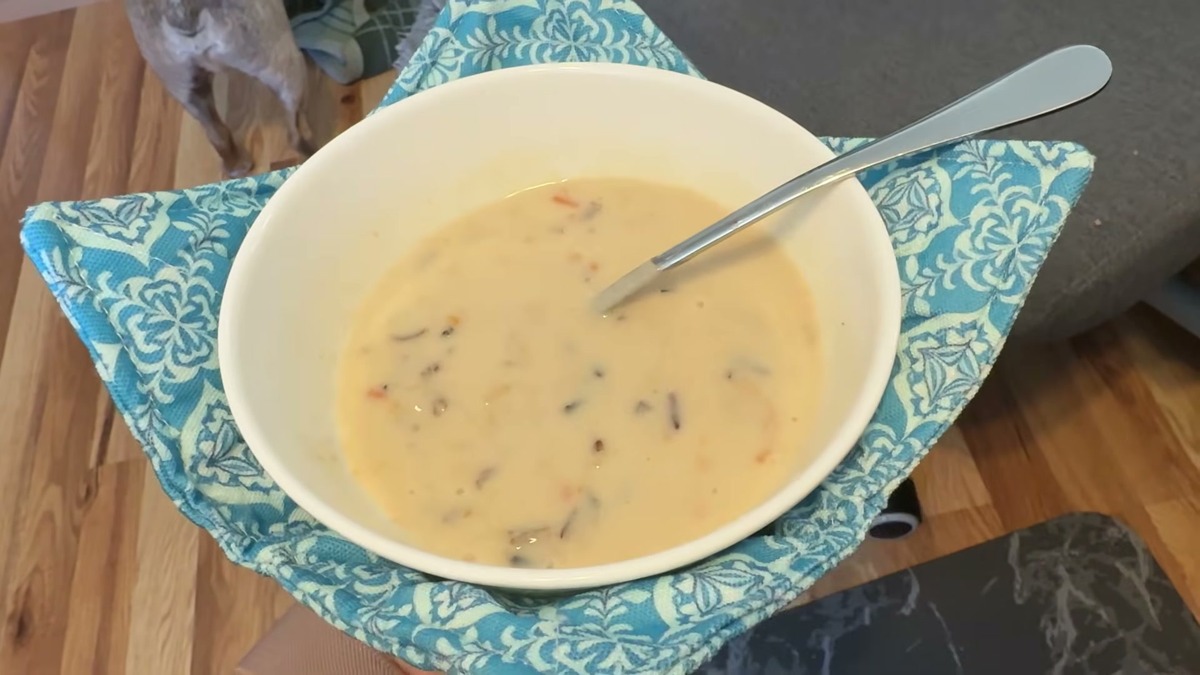 A white bowl of creamy soup with a spoon sitting in a teal patterned JounLoin cotton bowl cozy, held over a wood floor