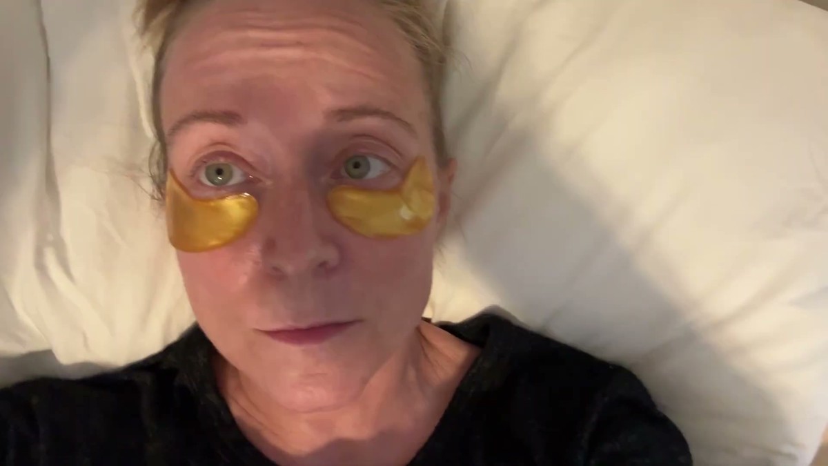 Jen lying back on a white pillow wearing a pair of gold 24K Maskiss hydrogel patches under her eyes