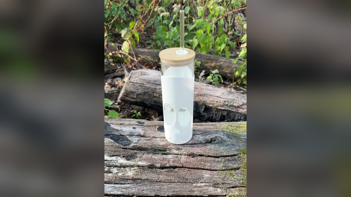 White skull-sleeved Bonesy glass tumbler with a bamboo lid and glass straw sitting on a weathered log in the woods