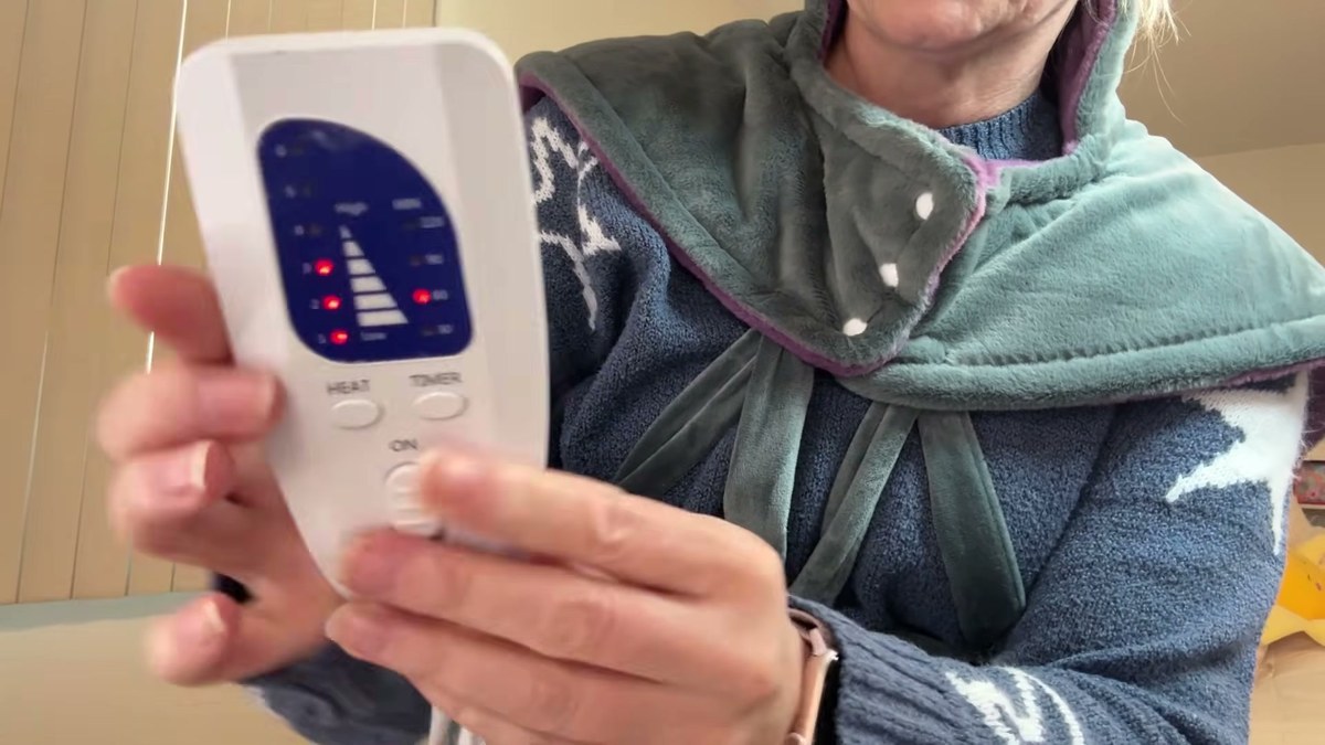 Hand holding the white reliefRX heating pad controller with red lights showing the heat level, next to the snapped high neck panel