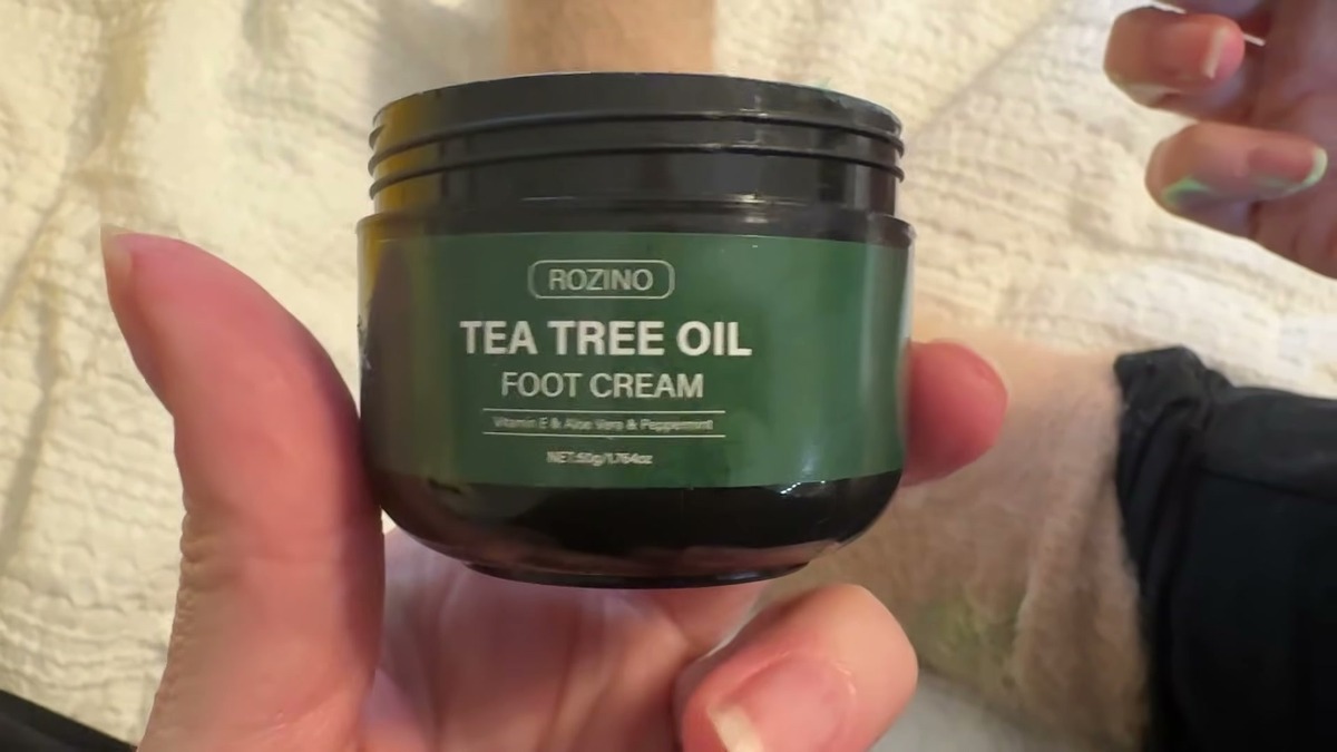 Hand holding an open black jar of Rozino Tea Tree Oil Foot Cream with a green label reading Vitamin E, Aloe Vera and Peppermint, 50g