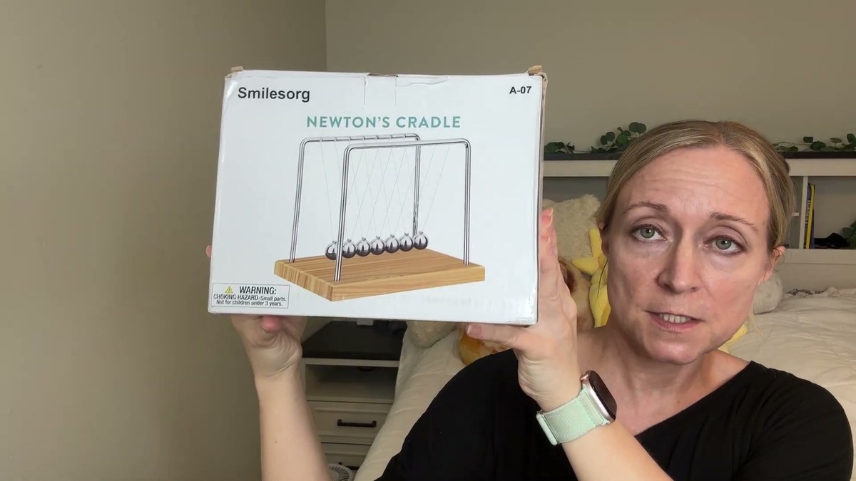 Jen holding up the white Smilesorg Newton's Cradle box, model A-07, with a picture of the cradle and a choking hazard warning